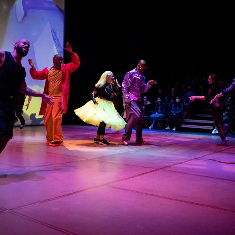 On a dancefloor, a woman with yellow hair and a yellow dress speaks into a microphone. Dancers run around her with their arms out.