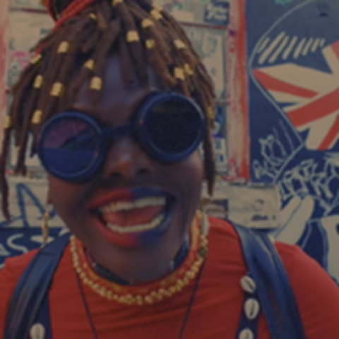 A Black woman with braids, and wearing goggles and red and blue lipstick on the left and right halves of her mouth, smiles wider.