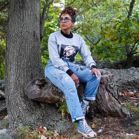 Sharinna sits on the base of a tree. She wears sunglasses and chuck taylors in the woods.