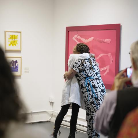 In a gallery, two women embrace with paintings on the wall behind them and a crowd clapping in the foreground.