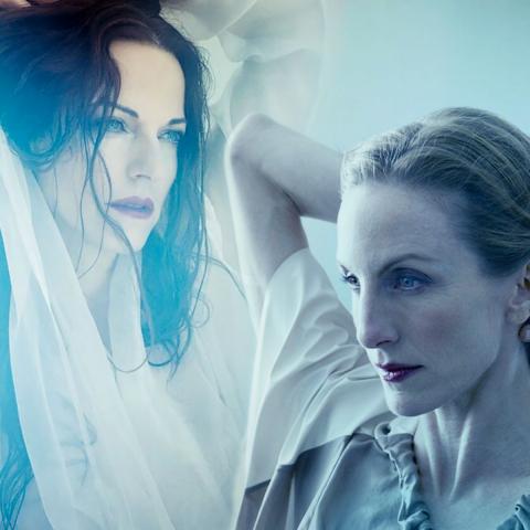 Two women face each other with one of their arms wrapped behind each of their heads.