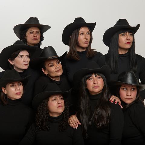 Nine women in black outfits and black cowboy hats pose like a family. They look as if they are seeing something off camera that makes them curious.