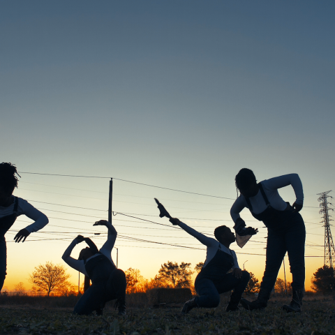 At sunset, four women, in overalls, dance in front of telephone lines.