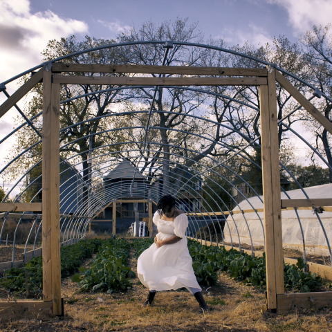 In front of the exposed strucutre of a greenhouse, a Black woman sits on a stool and looks down.