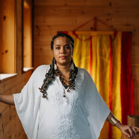 Iva has long brown hair. She is a woman of color and she wears white in a room with medium-stained wood.