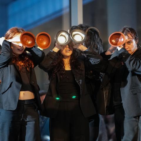 Three people dressed in black hold toilet plungers in front of their faces.