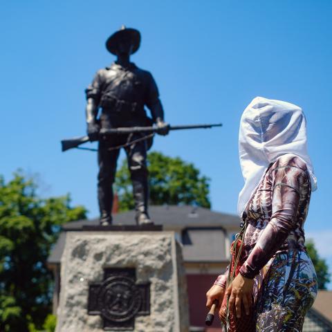 A statue of a white man holding a musket looms over a performer in a white shroud and body suit with a brown and white print.