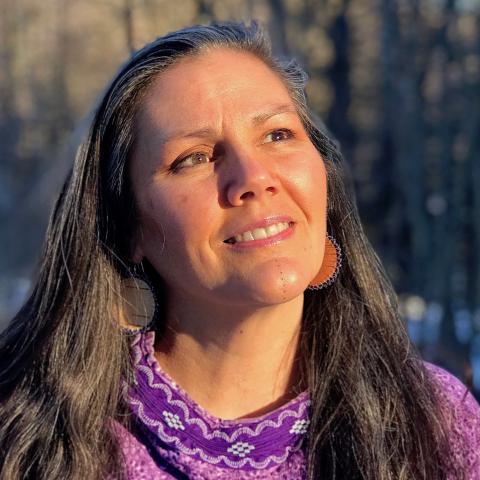 A woman with long dark hair smiles up into the sun. She is wearing a purple and white blouse and large round earrings. Trees and sunny sky are in the background.