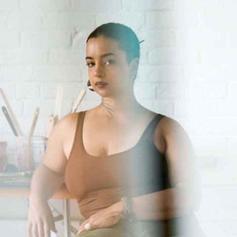 A woman with short dark hear wearing a brown tank top sits in a light room with a plant on a table in the background.