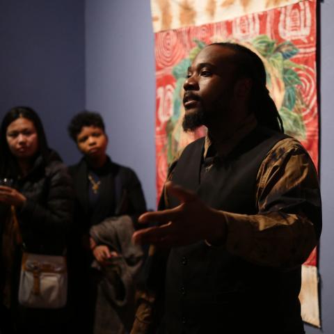 Stephen speaks to a room of people in front of a tapestry.