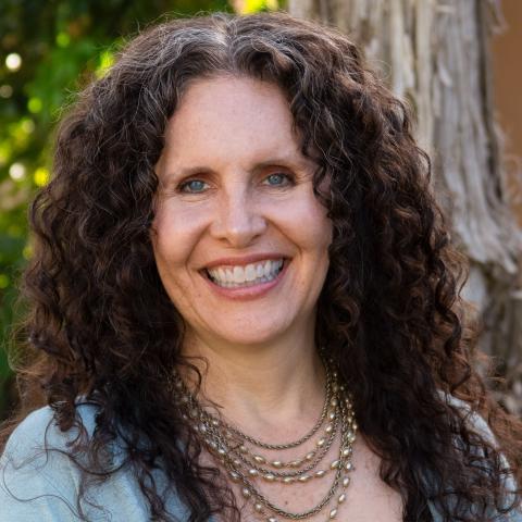 Shira has long, way brown hair. She wears multiple silver necklaces and a cardigan as she smiles next to a tree.
