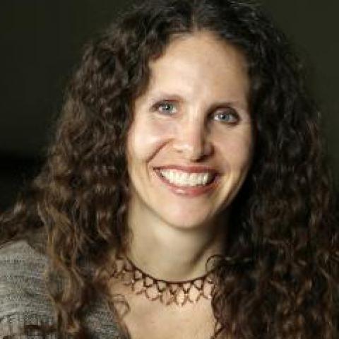 A woman with long curly hair smiles; she is wearing a gray sweater and a chocker necklace.