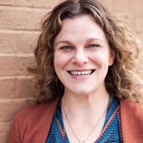 A head shot of a woman standing in front of a brick wall. She wears a blue patterned top under a rust color blazer. 