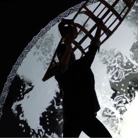 Silhouette of a figure in motion holding the skeleton of a birchbark canoe and a mask, the glow of illustrated waves behind them light up the canoe frame and shining through the eyes of the mask. 