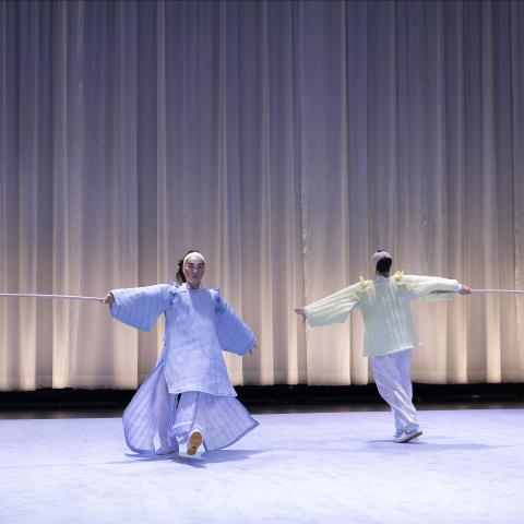 On a stage with a white curtain behind them, two performers hold white sticks with red strings at the end and face each other while walking in opposite directions, but possibly in a circle.