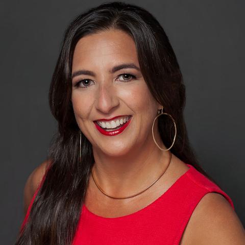 Stephanie smiles. She wears a red sleeveless dress and hoop earrings with red lipstick and long dark hair.