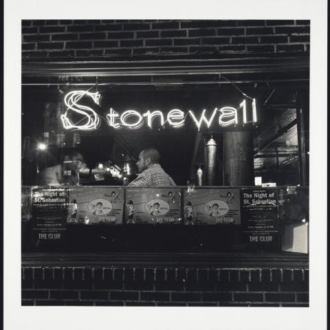 Outside, a window in a bar with the neon sign, "Stonewall Inn." A person sits on the other side.