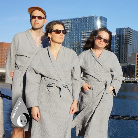 A man and two women in robes and sunglasses pose by the waterfront.