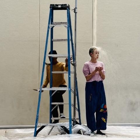 A woman stands behind a ladder while another woman has water dropped on her from an iv bag.