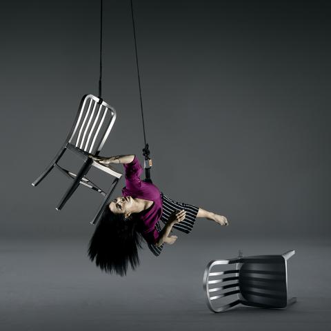 In a gray space, a performer glides while hanging next to a chair that is also strung from the ceiling. other chairs are turned over.