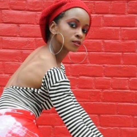 Tamara is a Black woman in a red beret.  She wears big hoop earrings and poses in front of bright red painted bricks.