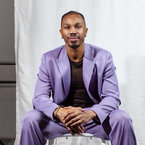 Tariq is a Black man. He wears a purple suit and mesh shirt in front of a white backdrop, with the room beyond the backdrop exposed.