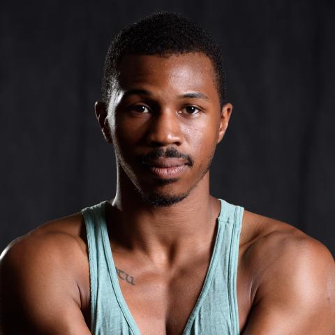 Tariq is a Black man with a mustache and a short hair cut. He wears a teal tank top in front of a black backdrop.