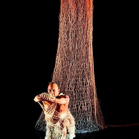 On a dark stage, a person stands in front of a net that drapes from the ceiling.