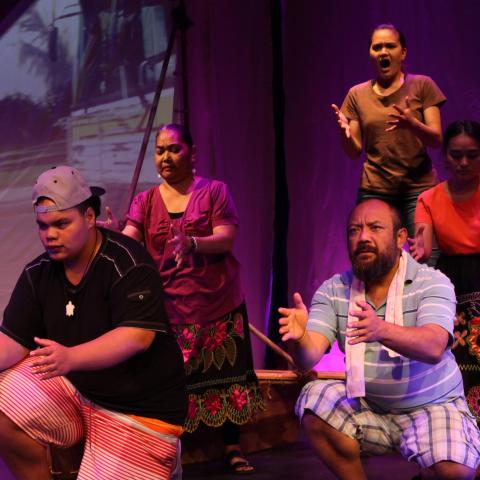 On a stage and next to a projection, five folks hold their arms out in front of them. Two in front, kneel, while three behind crouch. One sings.