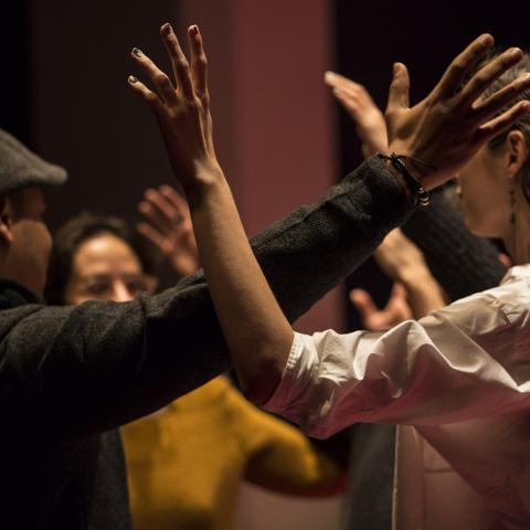 Two people are part of a larger crowd of people with their arms held up and touching each other at the elbows. The crowd is doing this in a circle.