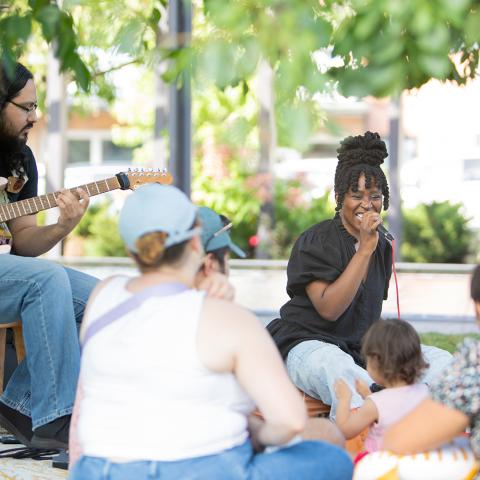 A Black woman sings into a microphone next to a guitarist. They're outside and in front of kids with chaperones.