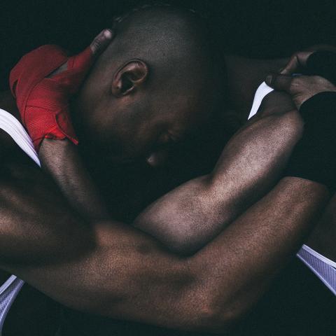 Two Black men in wrestling singlets and gloves hold each other in a head lock that looks almost like a hug.