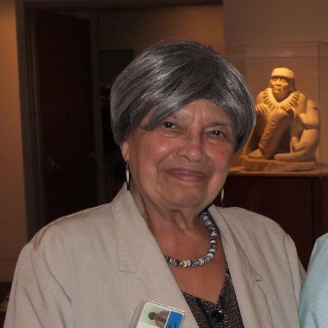 A woman with short gray hair wearing wampum earrings and necklace and a tan linen jacket; a figurine from the museum is in the background