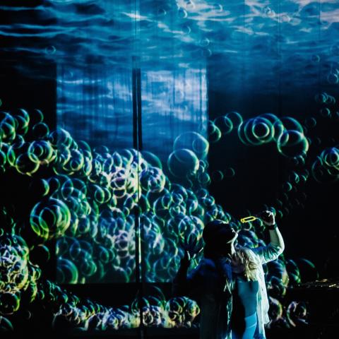 On a stage, a person blows lots of bubbles. Underlit, the bubbles create an oceanic print on the ceiling.