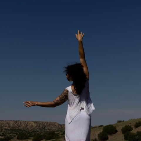 In the desert, a woman. with shoulder-length dark hair and in a white outfit, holds her right hand to the sky and left to the left.