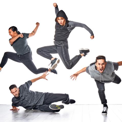 Four dancers, wearing gray, leap up, forward, or along the floor in front of a white backdrop.