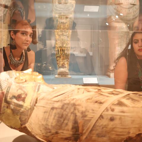 In a museum's glass case, a mummy. Outside the glass, two women peak inside.