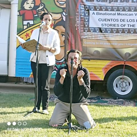 Outside, in front of a truck with a backdrop along it, four bandmates perform. The lead singer is on the ground on his knees with his hands in fists by his face.