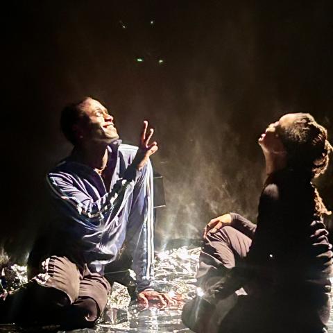 Two people sit on a square of tinfoil on a stage.