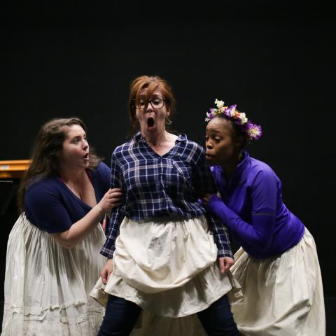 On a stage, three women in aprons argue.