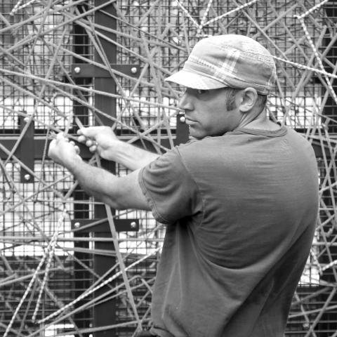 Black and white image: Randy holds some strings and looks back over his shoulder. He wears a hat and a t-shirt. He has light or white skin.