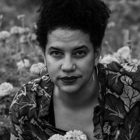 In black and white, Jessica, a light-skinned Black woman, makes eye contact. She wears floral and is outside.