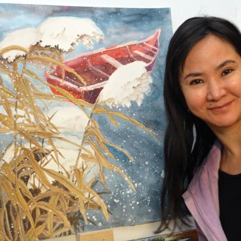 A young Chinese woman with long dark hair and wearing a lavender sweater smiles as she poses next to her painting  