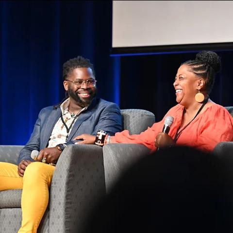 On a stage with three chairs, one facing another, three Black folks are in conversation and wear business attire.