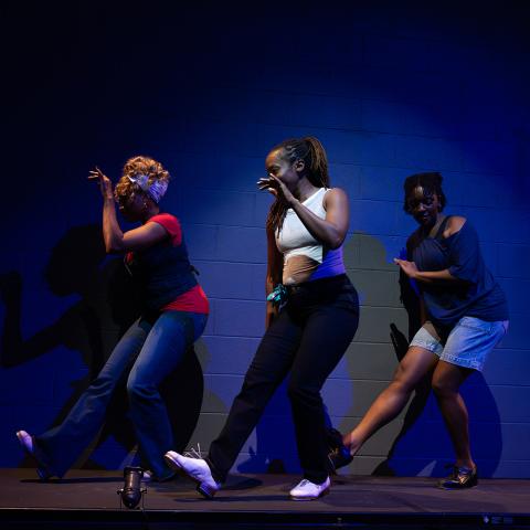 Three women perform in street clothes and in a spotlight. Behind them, a blue brick wall.