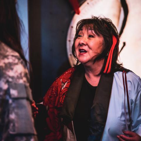 An elder Japanese woman speaks to another person. She stands in front of a gong and has a red ribbon tied to her hair.