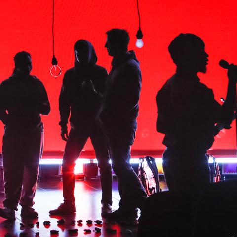 On a stage, with a red backdrop and lightbulbs hanging from the ceiling, four performer silhouettes sing into microphones.