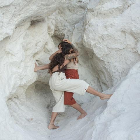 In a white rock formation, two women wrap their bodies around each other. They have brown hair and wear casual attire.