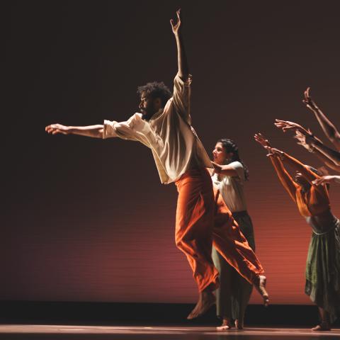 A male dancer leaps up while female dancers behind him reach towards him.