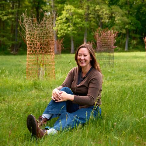 A woman with dark brown hair sits in a grassy field. Behind her, sculptures like chicken fence reach toward the sky.